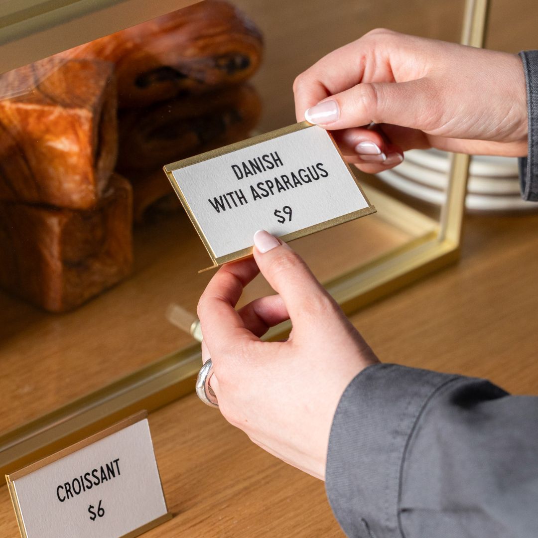 two hands holding brass card counter signs to place inside bakery display case