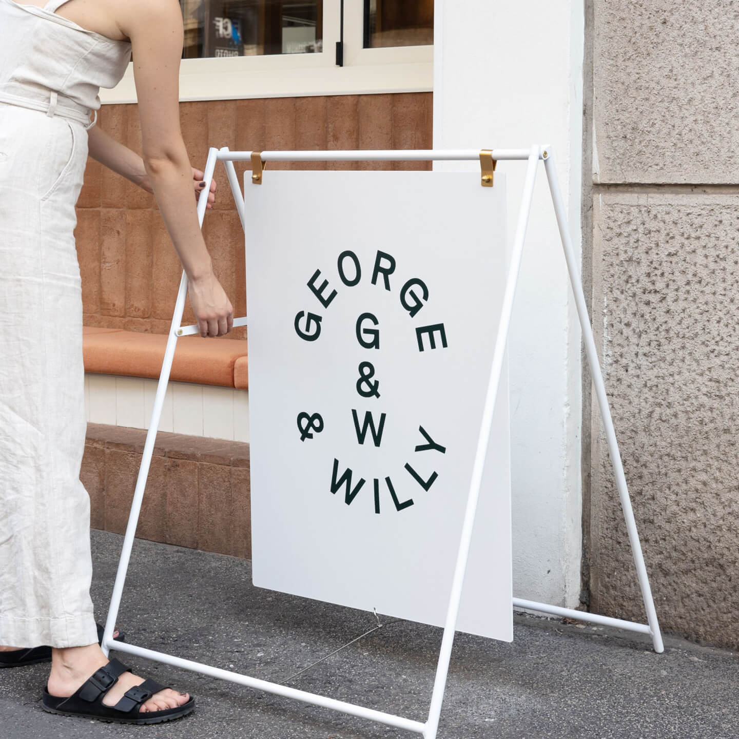 white large sidewalk pavement sign on sidewalk with george & willy logo with person adjusting the frame