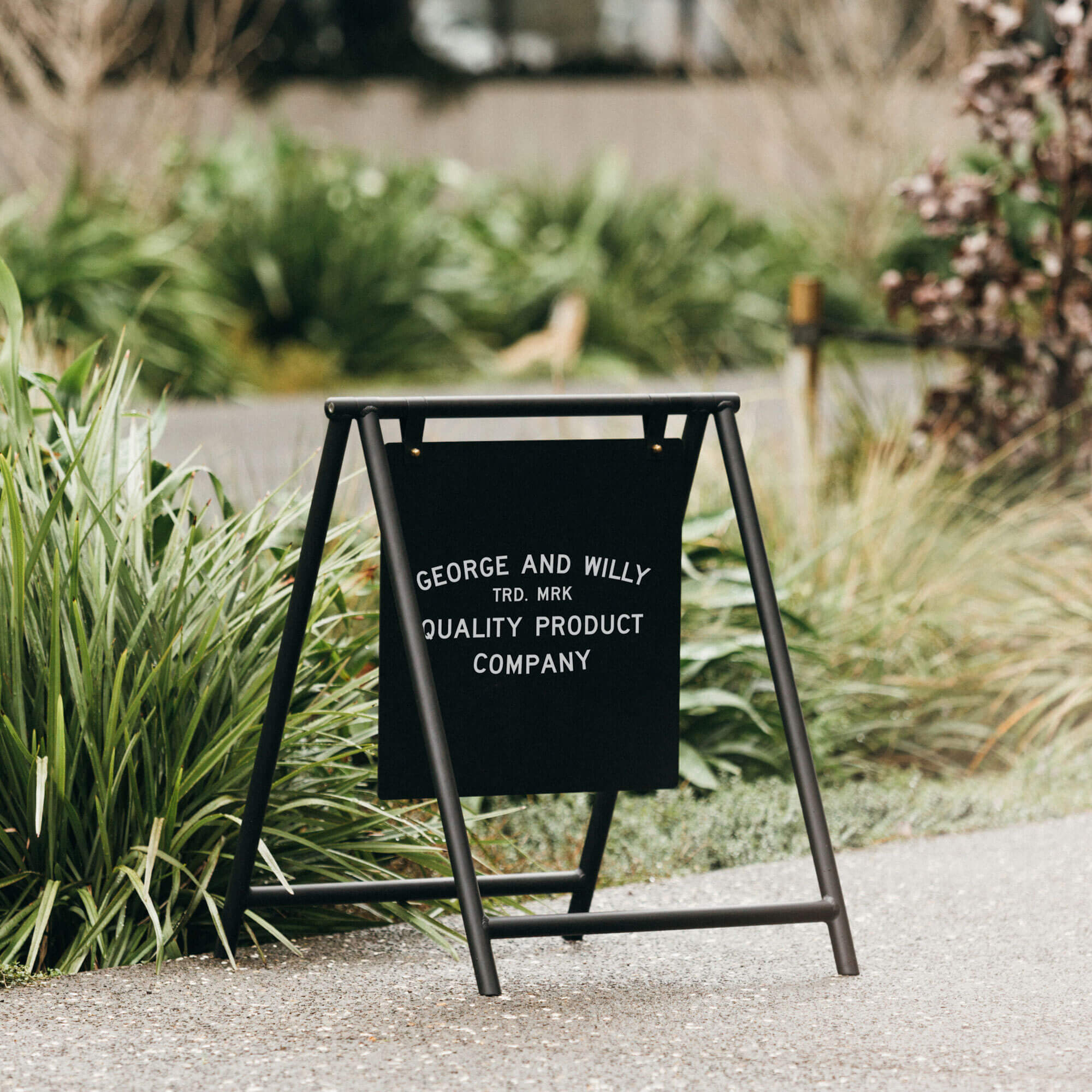Black A Frame sandwich board sign styled outdoors with shop logo message by plants