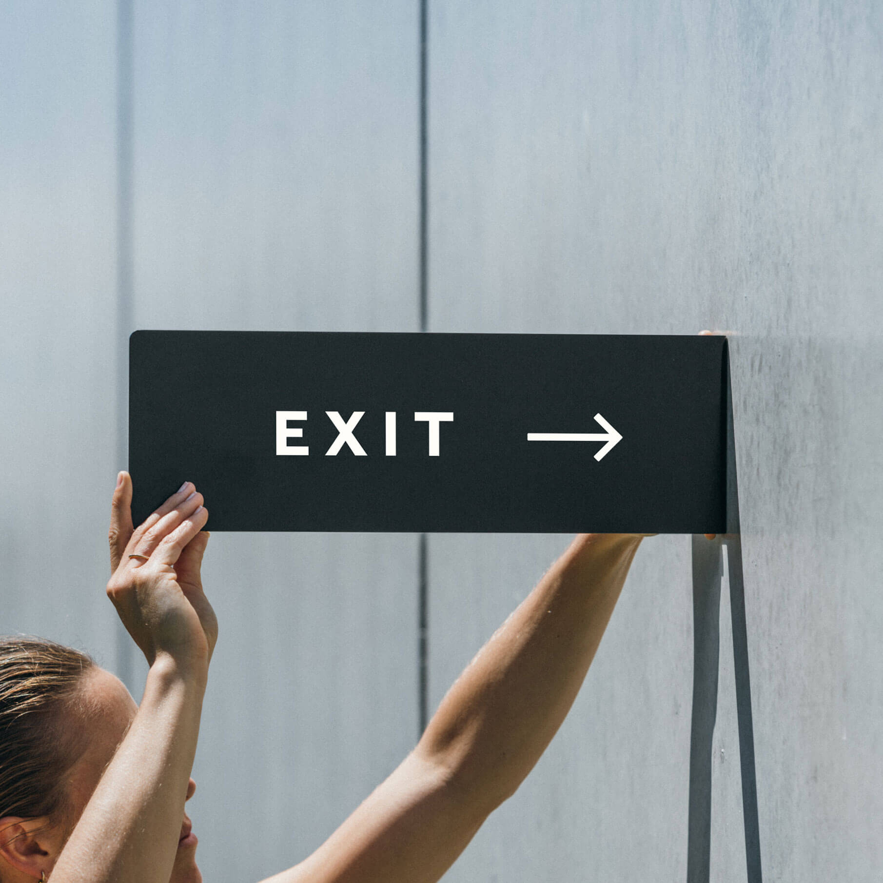 girl putting up the black rectangle blade sign on a cement wall wit the words "EXIT" on it