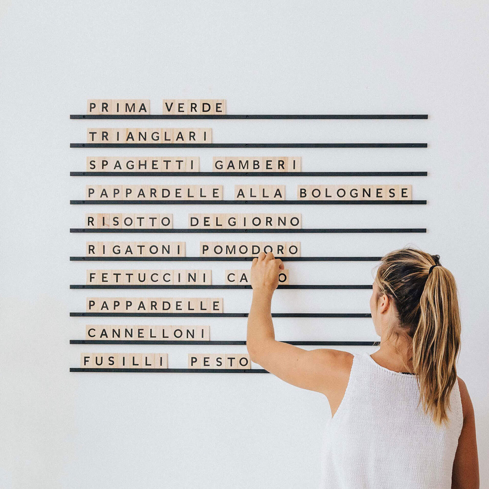 A girl adding wooden letter tiles to wall-mounted rails to display a pasta menu on a white wall