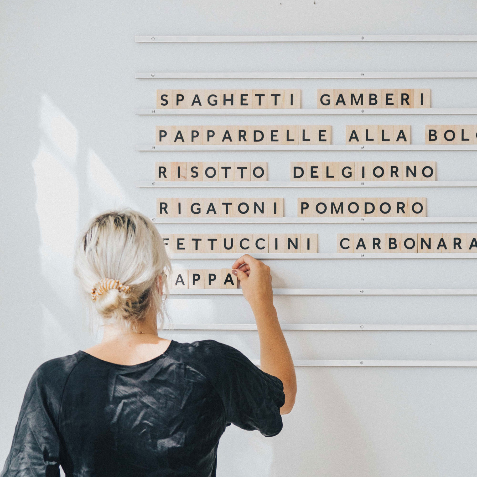 A girl adding wooden letter tiles to wall-mounted rails to display a pasta menu on a white wall